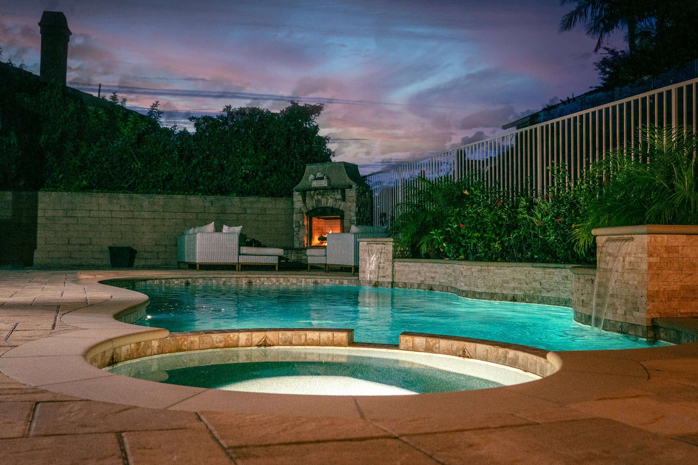 Custom backyard pool and spa with waterfalls and a lit fireplace at dusk